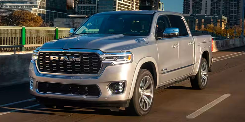 2025 Ram 1500 pickup truck driving on a city bridge in daylight, featuring a sleek silver exterior, bold front grille, and modern LED headlights with tall buildings in the background.