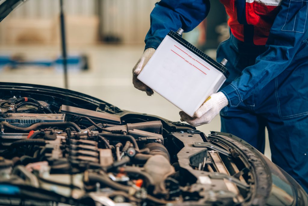 A mechanic wearing blue coveralls and white gloves installs a new car battery under the hood of a vehicle in an auto repair shop.